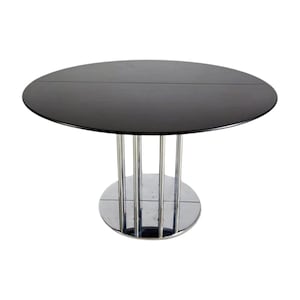 Könnte beinhalten: Ein moderner, runder Esstisch mit einer schwarzen Glasplatte. Der Tisch wird von einem Chromfuß mit vertikalen Metallstützen und einem kreisförmigen Sockel getragen. Das Design ist minimalistisch und elegant.