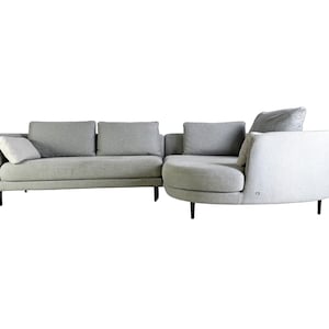Op de afbeelding: Een lichtgrijze hoekbank met een gebogen chaise longue. De bank heeft een modern ontwerp met zwarte poten en een zachte, comfortabele stof.