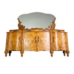 May include: A vintage wooden buffet with a curved front and three doors. The buffet has a mirrored top with a decorative carved frame. The wood has a rich brown color and a beautiful grain pattern.