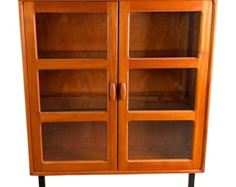 May include: A mid-century modern wooden cabinet with two glass-paned doors and two interior shelves. The cabinet is a warm, reddish-brown color and has black legs. The doors have small, rounded wooden handles.