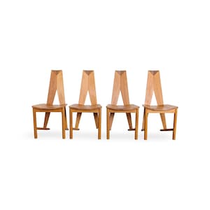 May include: A set of four light-toned wooden chairs, each featuring a tall, angular backrest with a V-shaped central cutout and a flat, contoured seat. The chairs are supported by four straight legs, with two angled rear legs extending upward.