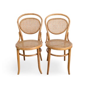 May include: A pair of light brown wooden chairs featuring curved frames and circular backrests with woven cane inserts. Each chair has a round seat also finished with a woven cane pattern, supported by four slender, slightly flared wooden legs.