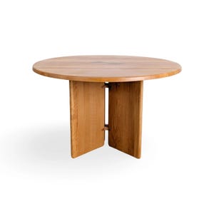 May include: A round wooden dining table featuring a smooth, light brown surface with a visible grain pattern. The table is supported by a central base composed of two vertical panels joined together by small, dark, rectangular wooden connectors.