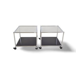 May include: Two modern side tables. Each table has a clear glass top, a black lower shelf, and a chrome frame. The tables are on small wheels for easy movement. The design is minimalist and functional.