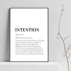 Op de afbeelding: Een zwart omkaderde print met het woord "INTENTION" in zwarte letters. Daaronder staat de definitie van het woord. De print wordt gepresenteerd op een witte ondergrond, met een takje witte bloemen op de achtergrond. De tekst is duidelijk en leesbaar.