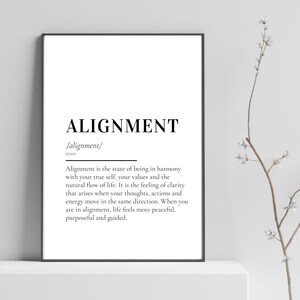 Op de afbeelding: Een zwart-wit print met het woord "ALIGNMENT" in een vetgedrukte lettertype en de definitie. De print wordt gepresenteerd op een wit oppervlak. Rechts van de print bevindt zich een tak met witte bloemen.