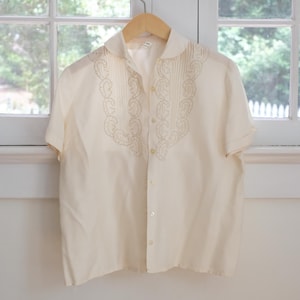 May include: A short-sleeved cream-colored button-down shirt featuring a pointed collar and vertical pleats on the front chest. The shirt is decorated with intricate tan embroidery in a swirling pattern along the pleated panels and has buttons.