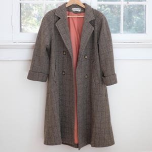 May include: A double-breasted brown coat featuring a subtle dark grid pattern, wide lapels, and cuffed sleeves. The garment is fully lined with a solid salmon-colored fabric and includes four visible buttons on the front closure.