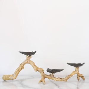 May include: A decorative, three-tiered serving stand. The stand features a natural, branch-like design with a gold and beige finish. Three dark gray, leaf-shaped dishes are attached to the branches. The stand is placed on a white marble surface.