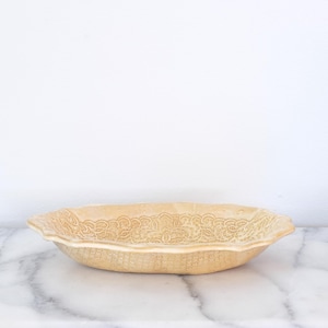 May include: A beige ceramic oval serving dish with a detailed floral lace pattern around the rim. The dish has a scalloped edge and is set against a white background.
