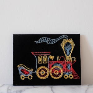 May include: A black velvet framed artwork depicting a colorful string art train with a blue smoke plume. The train is red, yellow, and blue with a yellow caboose.