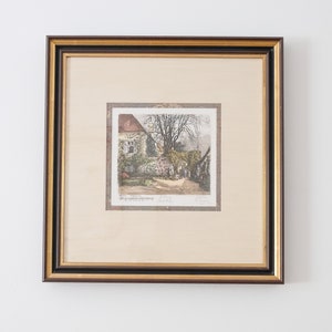 May include: A framed print of a vintage illustration depicting a quaint cottage with a lush garden and a winding path. The print is in a brown frame with a gold trim.