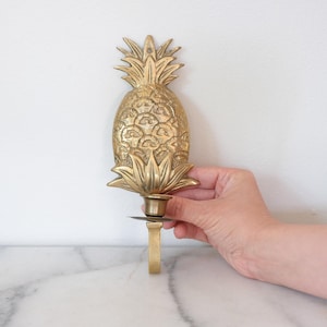 May include: A brass pineapple-shaped wall sconce. The sconce features detailed leaf and fruit textures, with a candle holder at the base. The pineapple is a warm gold color, and the sconce is designed to be mounted on a wall.