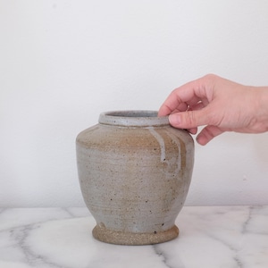 May include: A handcrafted, speckled ceramic vase with a wide, rounded body and a slightly flared rim. The vase has a neutral color palette with shades of gray and brown, and a textured surface. The base is a darker shade of brown.