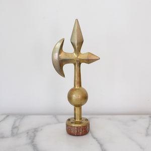 May include: A brass decorative object featuring a central vertical shaft topped with a three-pointed head, including a curved blade, a pointed top, and a pointed side. A spherical brass element sits above a circular base with a textured wood finish.