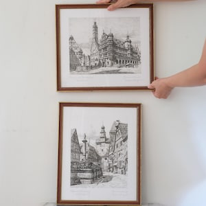 May include: Two framed black and white architectural etchings depicting historic town scenes with detailed buildings, towers, and fountains. Each print is encased in a thin brown wooden frame with gold trim, showcasing intricate line work and shading.