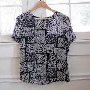 May include: A short-sleeved blouse with a patchwork design featuring zebra and leopard prints in black and white. The top is hanging on a wooden hanger in front of a window.
