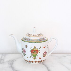 May include: White porcelain teapot with a lid, spout, and handle. The teapot is decorated with colorful floral and ornamental designs, including a central vase motif. The lid and base feature intricate patterns with gold accents. A classic piece for tea lovers.