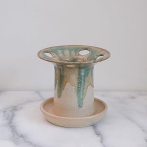 May include: A ceramic flower frog with a wide, flared top featuring multiple circular openings. The piece has a cream-colored base with a glossy, teal-green glaze dripping down from the rim. It sits on a matching circular ceramic saucer base.