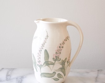 May include: A white ceramic pitcher with a floral design. The pitcher has a wide mouth and a handle. The floral design features pink and green flowers.