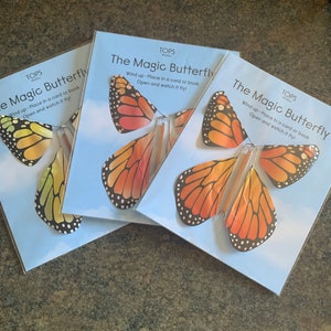 Op de afbeelding: Drie pakjes met het opwindbare speelgoed "The Magic Butterfly". Elk pakje heeft een kleurrijke vlinder met oranje, gele en zwarte vleugels. De tekst op het pakje luidt "TOPS Malibu The Magic Butterfly Wind up - Place in a card or book Open and watch it fly!"