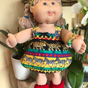 May include: A Cabbage Patch Kid doll wearing a yellow, green, and black dress with a zebra and leopard print. The doll has blonde hair, blue eyes, and is wearing red crocheted shoes.