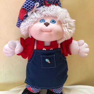 May include: A white plush doll with blue eyes and a brown nose, wearing a red shirt, blue overalls, and a blue and white polka dot hat with strawberries. The doll is standing on a white surface.