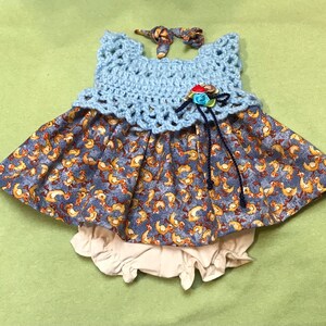 May include: A blue and white crocheted dress with a floral embellishment. The dress has a skirt with a pattern of yellow ducks on a blue background. The dress is paired with a white crocheted diaper cover.