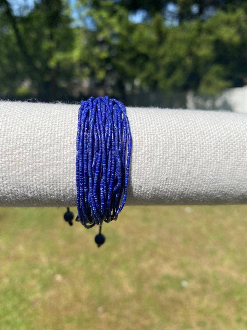 May include: A blue beaded bracelet with black cord ties. The bracelet is made of small, round beads and is wrapped around a white fabric surface.