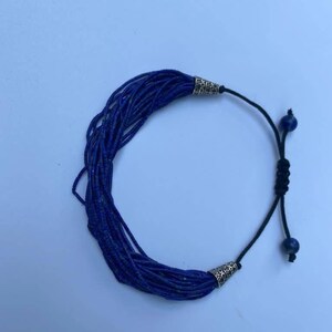 May include: A blue lapis lazuli beaded bracelet with silver end caps and a black cord with a sliding knot closure. The bracelet features two small lapis lazuli beads as accents.