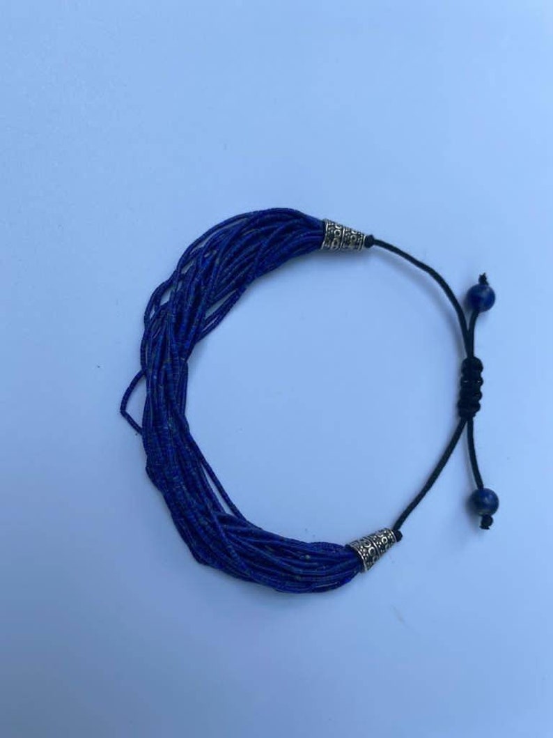 May include: A blue lapis lazuli beaded bracelet with silver end caps and a black cord with a sliding knot closure. The bracelet features two small lapis lazuli beads as accents.