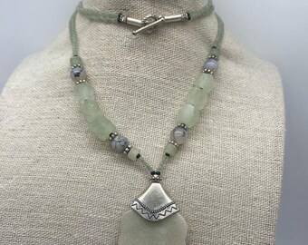 May include: A green and silver necklace with a large, leaf-shaped pendant. The necklace is made of various green and grey beads, and the pendant has a silver design.