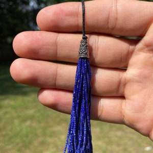 May include: A blue lapis lazuli tassel with a silver-toned bead at the top. The tassel is hanging from a black cord.