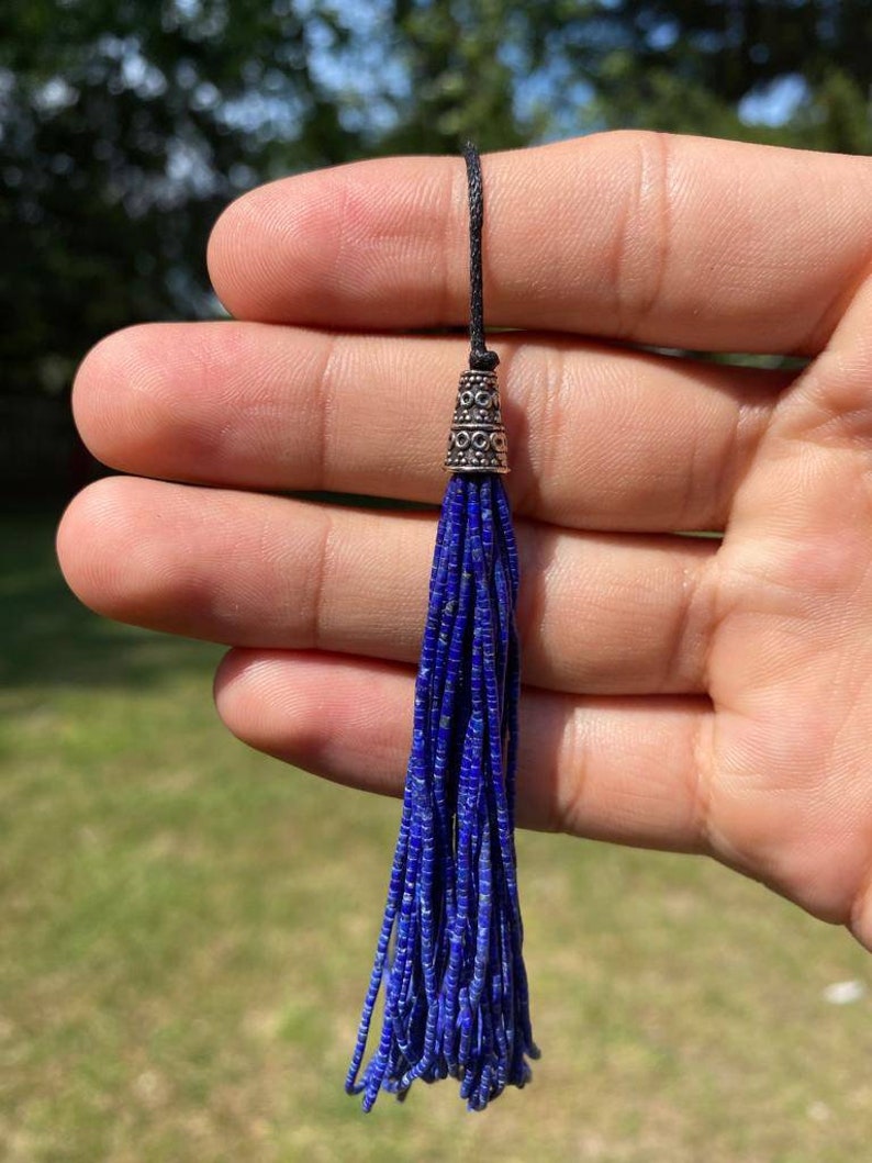 May include: A blue lapis lazuli tassel with a silver-toned bead at the top. The tassel is hanging from a black cord.
