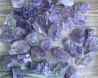 May include: A collection of raw amethyst crystals in various sizes and shapes. The crystals are a deep purple color and have a natural, rough texture. They are arranged on a wooden surface.