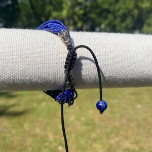 May include: A blue and black bracelet with silver accents and two lapis lazuli beads. The bracelet is made of multiple strands of blue cord and has a tassel at the end.