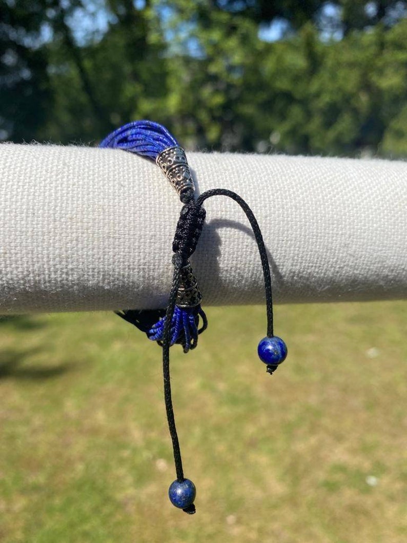 May include: A blue and black bracelet with silver accents and two lapis lazuli beads. The bracelet is made of multiple strands of blue cord and has a tassel at the end.
