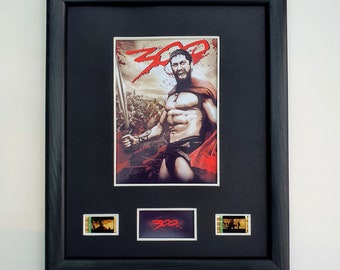 May include: Framed movie poster for the film "300" featuring a muscular warrior holding a sword, with the title in red. The black frame includes three smaller images below the main poster.