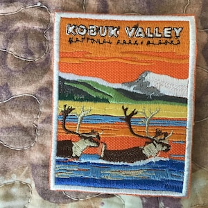 May include: A rectangular embroidered patch featuring a landscape of Kobuk Valley National Park, Alaska. The design shows two caribou wading through blue water, with green hills, a snow-capped mountain, and an orange sky. Text reads KOBUK VALLEY.