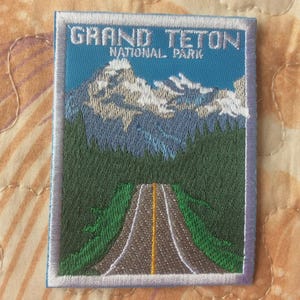 Puede incluir: Parche bordado con una vista del Parque Nacional Grand Teton. El diseño incluye una carretera que conduce a una cordillera con picos nevados, sobre un cielo azul. El parche está bordeado en blanco y azul.