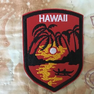 Pode incluir: Um emblema bordado em forma de escudo com fundo vermelho e borda preta. O design apresenta o texto branco HAWAII no topo, silhuetas de palmeiras pretas, um sol amarelo e um pequeno barco preto na água na parte inferior do emblema.