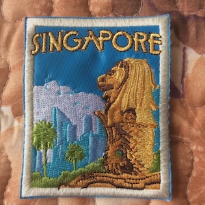 Könnte beinhalten: Gestickter Aufnäher mit dem Wort "SINGAPORE" in goldfarbenen Buchstaben. Er zeigt den Merlion, die Skyline von Singapur und Palmen auf blauem Hintergrund mit weißem Rand. Rechteckige Form.