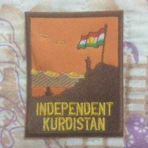 Può includere: Toppa ricamata con una scena paesaggistica, una bandiera e la scritta "INDEPENDENT KURDISTAN". La toppa ha un bordo marrone, un cielo arancione e una bandiera con colori rosso, bianco, verde e giallo. Il design include sagome di figure e montagne.