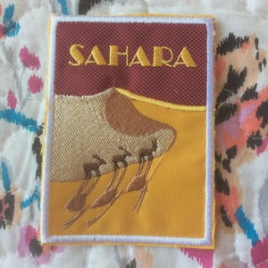 May include: Embroidered patch with the word "SAHARA" in gold thread. The design shows a desert scene with a sand dune, three camels, and a yellow and brown colour scheme. The patch has a white border.