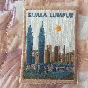 May include: Embroidered patch featuring the Kuala Lumpur skyline, including the Petronas Towers. The patch has a beige border and the text "KUALA LUMPUR" at the top. The design includes blue, brown, and yellow elements.