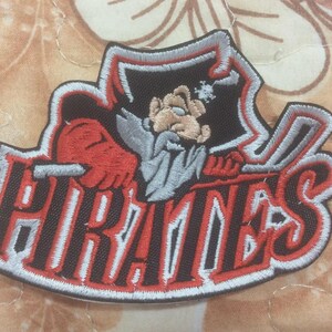Könnte beinhalten: Gestickter Aufnäher mit dem Wort "PIRATES" in Rot und Weiß, umrandet in Schwarz. Er zeigt eine Cartoon-Piratenfigur mit roter Hand und grauem Bart. Der Aufnäher hat einen weißen Rand.
