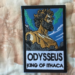 Puede incluir: Un parche rectangular bordado que muestra una figura de piel dorada con barba oscura y cabello rizado sobre un cielo azul y un paisaje verde. Debajo de la figura, una sección blanca contiene el texto en negro ODYSSEUS KING OF ITHACA.