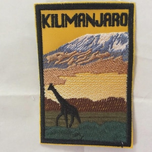 以下が含まれることがあります： 「KILIMANJARO」の文字が入った刺繍ワッペン。山脈、キリンのシルエット、黄色、茶色、緑色の風景がデザインされています。ワッペンは黒い縁取りがされています。