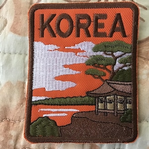 May include: Embroidered patch featuring the word "KOREA" in brown letters above a scenic landscape. The design includes a traditional building, trees, and a body of water, all set against an orange and white sky.