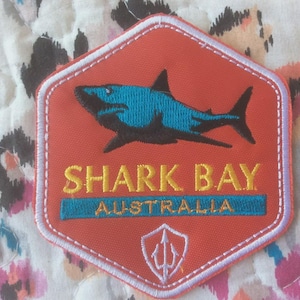 Puede incluir: Parche bordado con un tiburón azul sobre las palabras "SHARK BAY AUSTRALIA". El parche tiene forma hexagonal con borde blanco y un símbolo de tridente. Fondo floral.